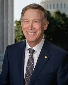 Official portrait of John Hickenlooper, U.S. Senator from Colorado, wearing a suit and tie, smiling in front of a blurred background.