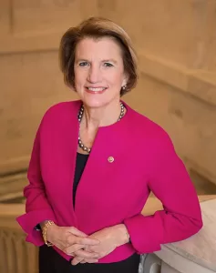 Official portrait of Shelley Moore Capito, U.S. Senator from West Virginia, wearing a bright pink jacket and smiling in front of a marble background.