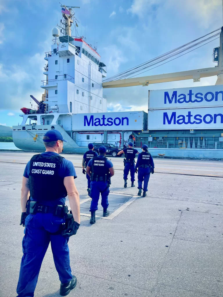 United States Coast Guard personnel conducting a security inspection at a port, walking towards a Matson cargo ship under a partly cloudy sky. This is part of their mission which also includes maritime cybersecurity