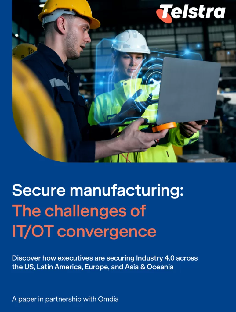 Workers in a manufacturing facility discuss IT-OT security in manufacturing while using a laptop. A digital interface overlay illustrates advanced technology integration. Telstra logo appears in the top right corner. The text on the image highlights the challenges of IT/OT convergence in securing Industry 4.0.