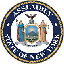 The image is the official seal of the New York State Assembly. It features a circular design with a blue outer ring containing the words "ASSEMBLY" at the top and "STATE OF NEW YORK" at the bottom in bold, capitalized white letters. The inner part of the seal displays the New York State Coat of Arms, which includes a shield with a sun rising behind a mountain and river, supported by two allegorical figures: Liberty (holding a pole with a Phrygian cap) on the left and Justice (holding scales and a sword) on the right. Above the shield, a bald eagle sits atop a globe. Below the shield, a banner displays the state motto, "Excelsior", meaning "Ever Upward."