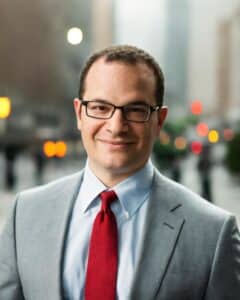 eMazzanti Technologies President Carl Mazzanti  in a suit and red tie smiling outdoors in a city setting, used in an article about holiday scams and cybersecurity awareness.